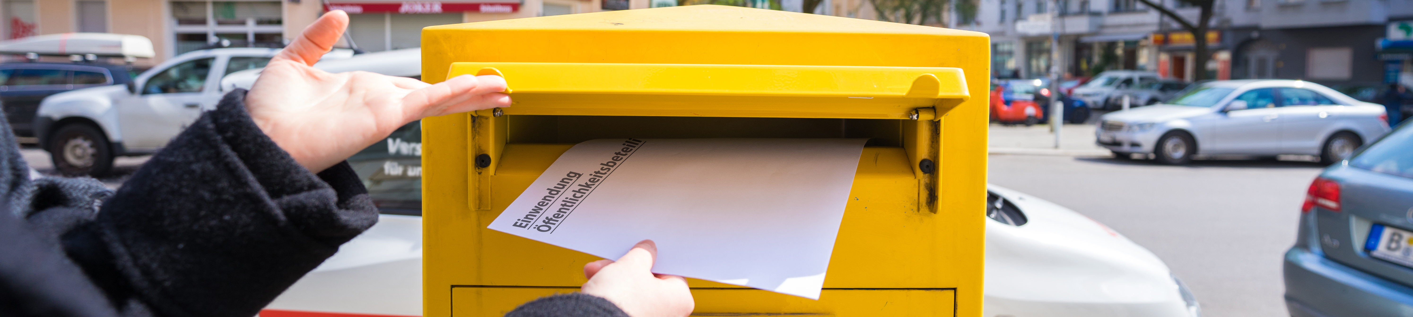 Einwurf eines Umschlags mit den Schlagwörtern Einwendung und Öffentlichkeitsbeteiligung in Postbriefkasten  